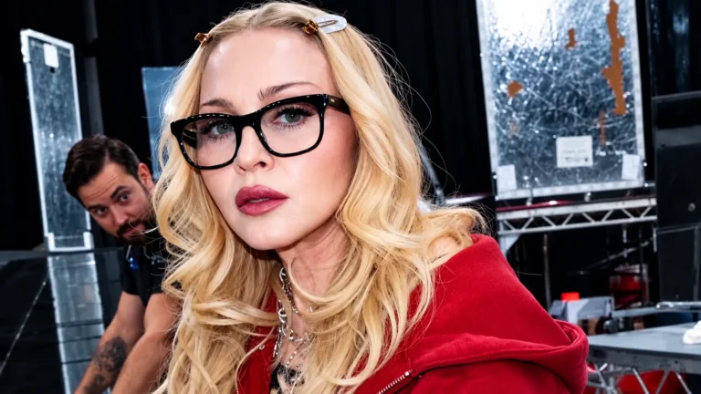 Madonna com cabelos loiros, óculos pretos e figurino vermelho em imagem editorial inspirada nos bastidores do VMA 2026.