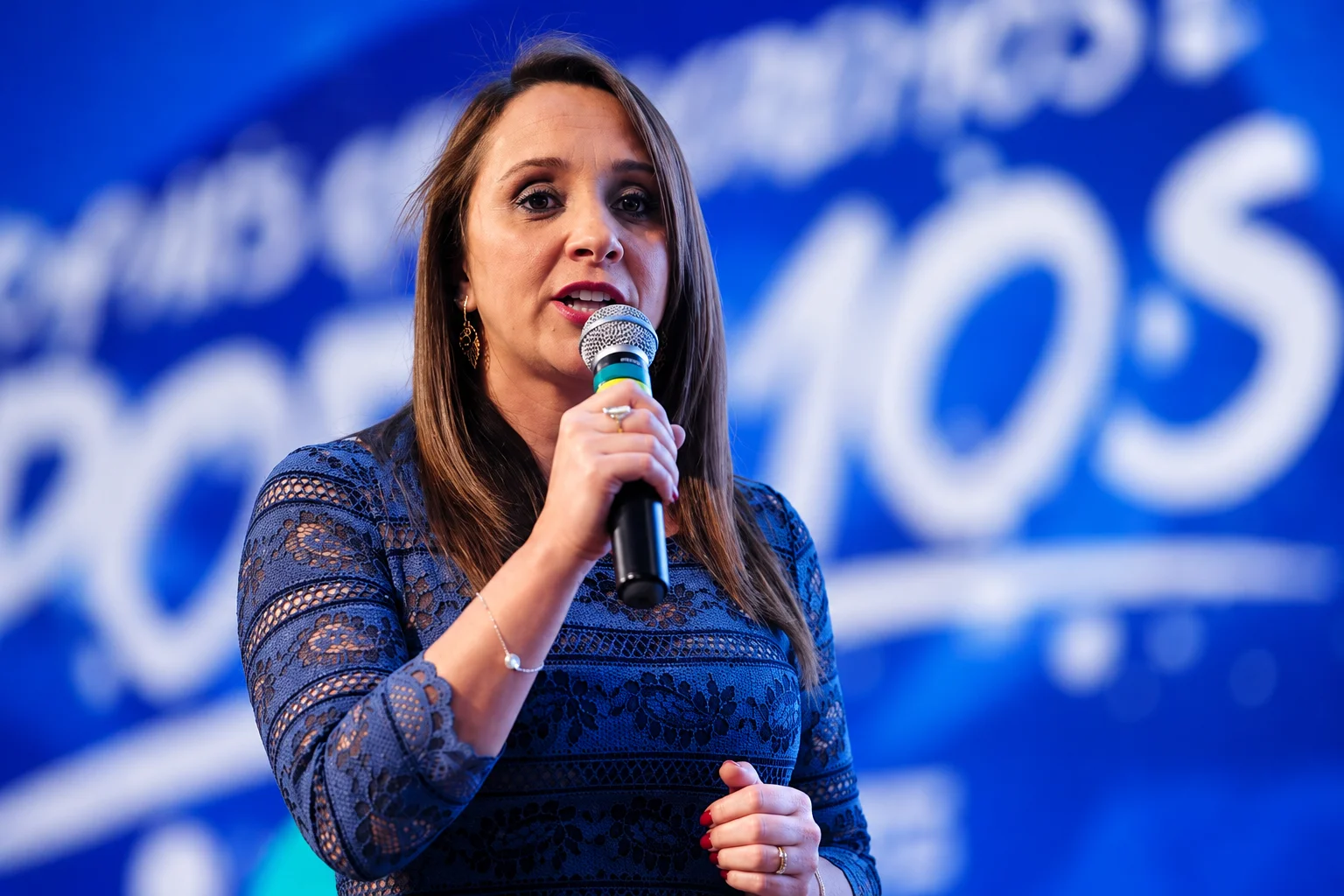 Mulher apresentada em reportagem sobre Thiago Ganem discursando ao microfone durante evento político